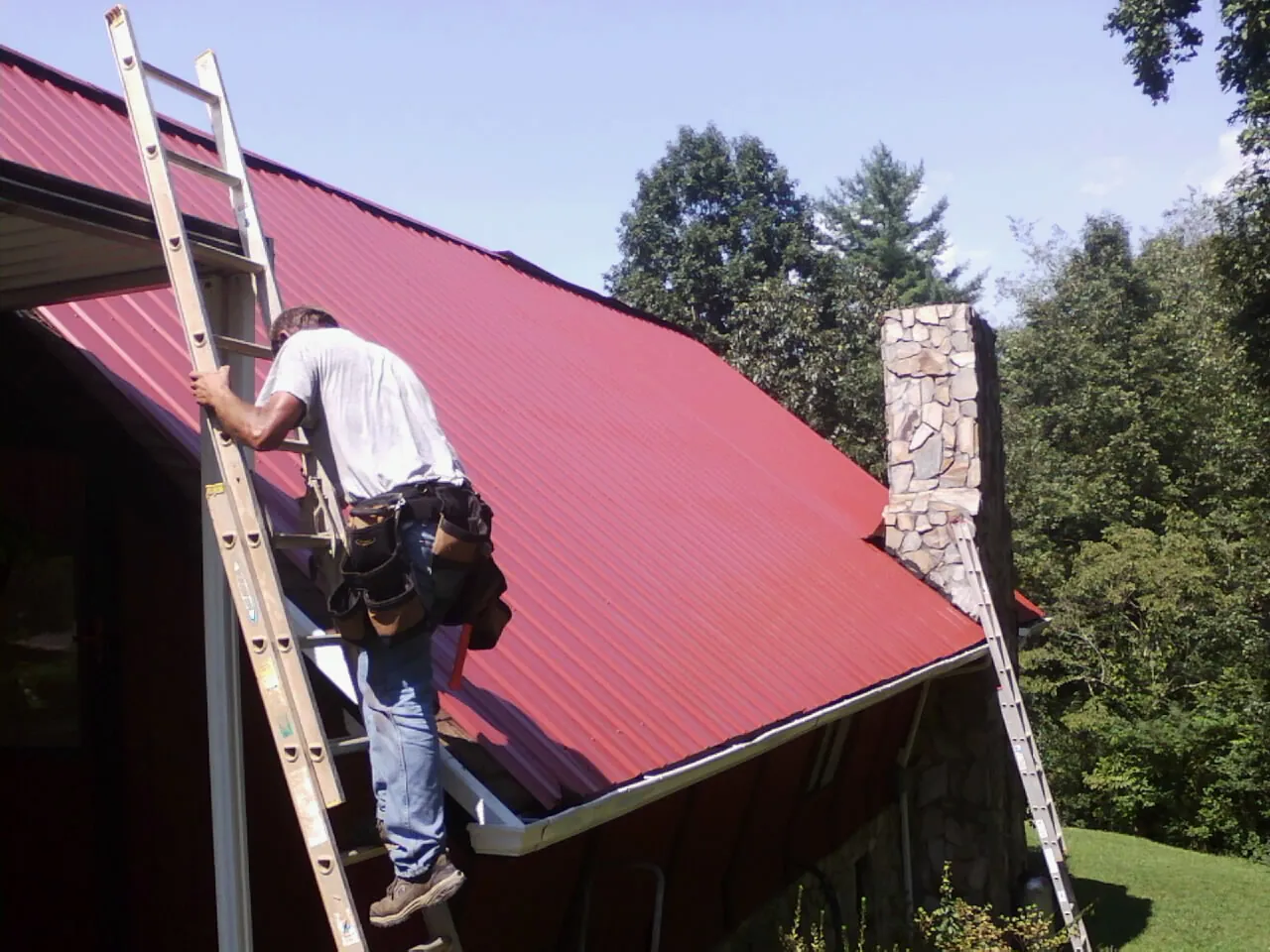Roofer on a ladder beside a red metal roof and stone chimney — Soffit & Fascia Repair by Roofing Brothers in Pescadero